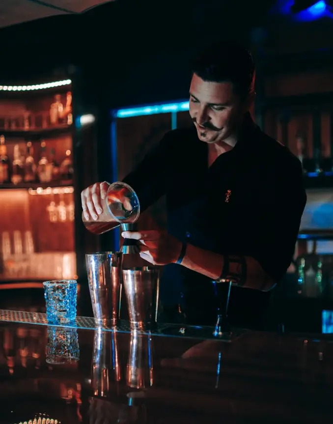 Bartender pouring a drink behind a bar, focused on mixing a cocktail under warm, moody lighting.