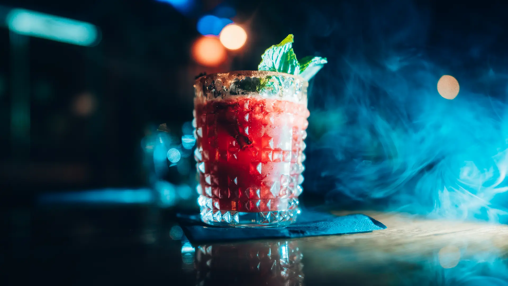 Red cocktail in a textured glass with a garnish, sitting on a bar surface surrounded by blue-tinted smoke.