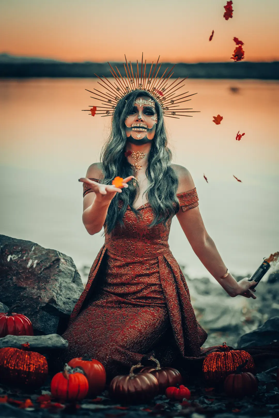 Person in a red dress with dramatic face paint and a spiked headpiece, standing outdoors with pumpkins and falling leaves.