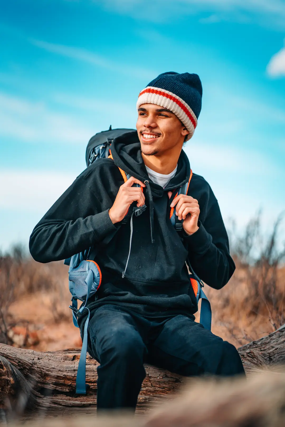 Person wearing outdoor apparel and a backpack, smiling while seated on a log in a natural landscape.