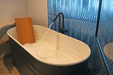 Black and white bathtub filling with water and foam, with a wooden board resting on its edge in a modern bathroom.
