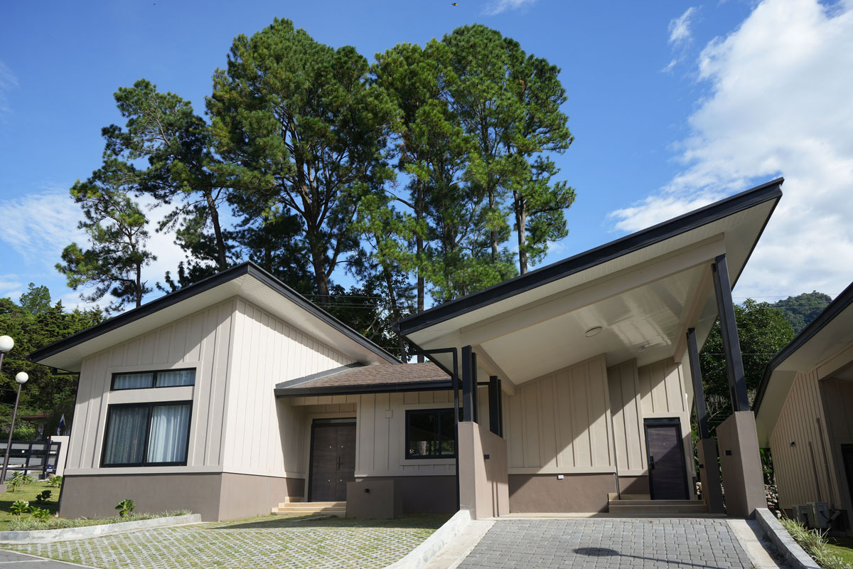 Casa moderna de un solo piso con techo inclinado, rodeada de árboles altos bajo un cielo azul con algunas nubes.