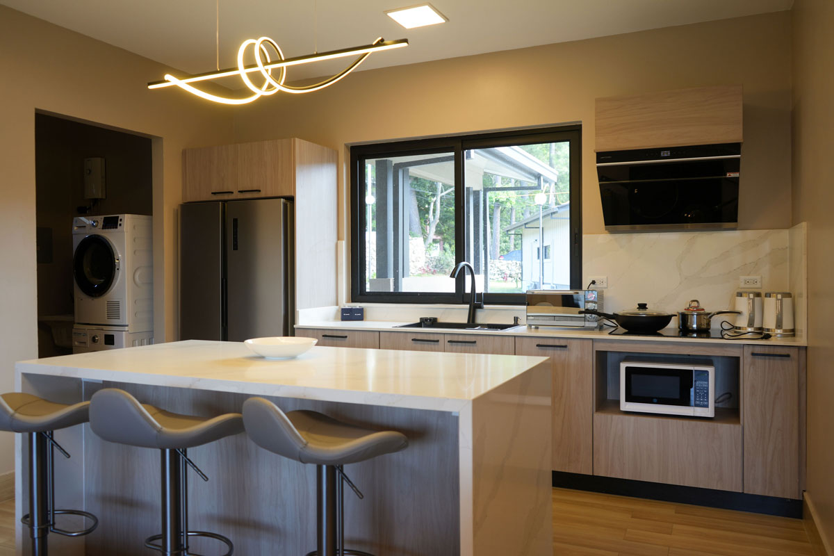 Cocina moderna con isla central blanca, tres taburetes grises, electrodomésticos de acero inoxidable, ventana amplia y lámpara colgante artística.