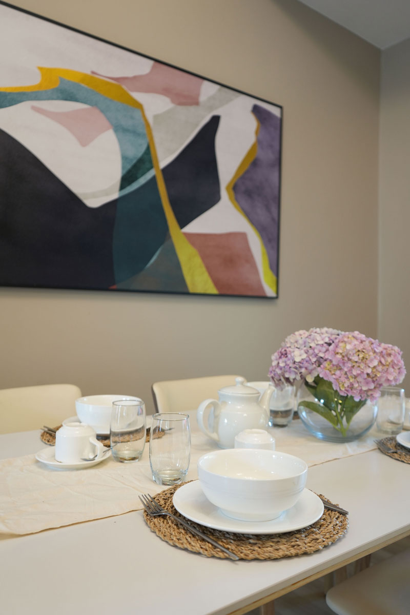 Mesa de comedor con vajilla blanca, vasos de cristal, tetera y un arreglo floral de hortensias lilas en un jarrón transparente.