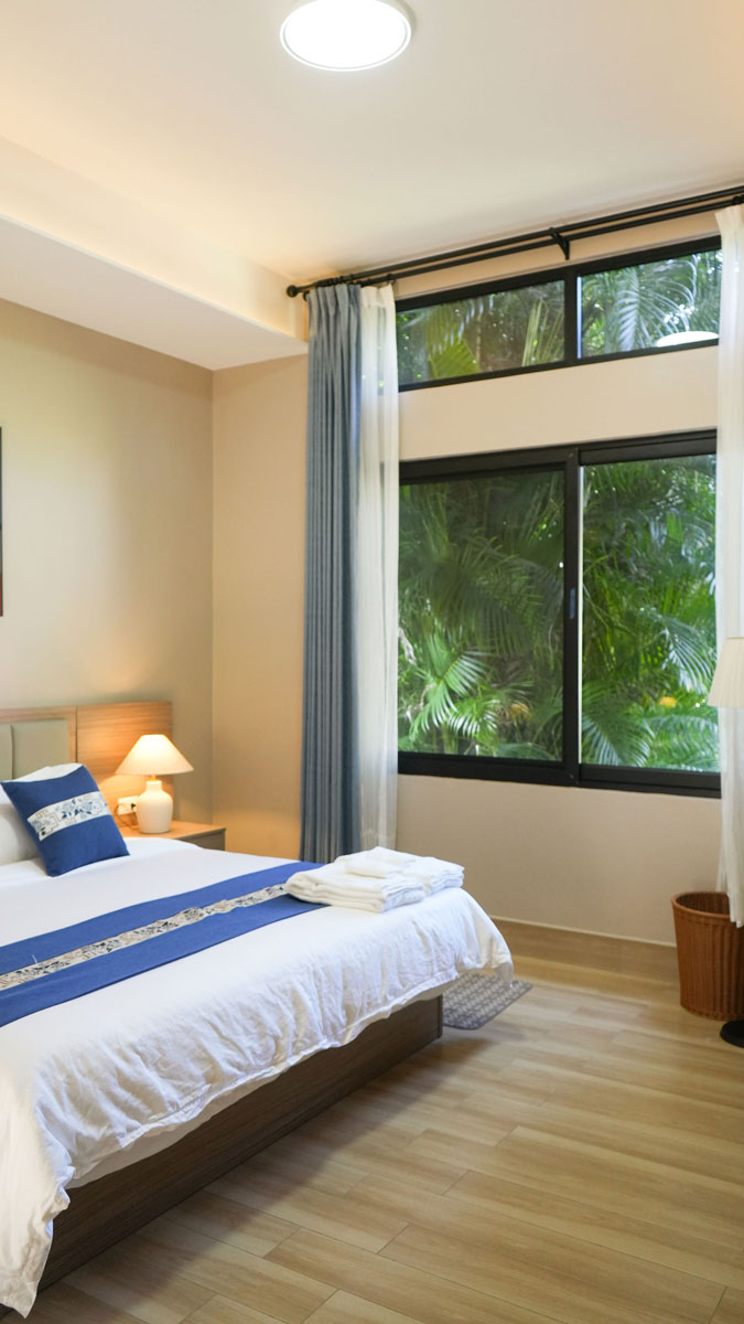 Dormitorio con cama doble, ropa de cama blanca y azul, lámpara de noche encendida y ventana grande con vista a plantas verdes.