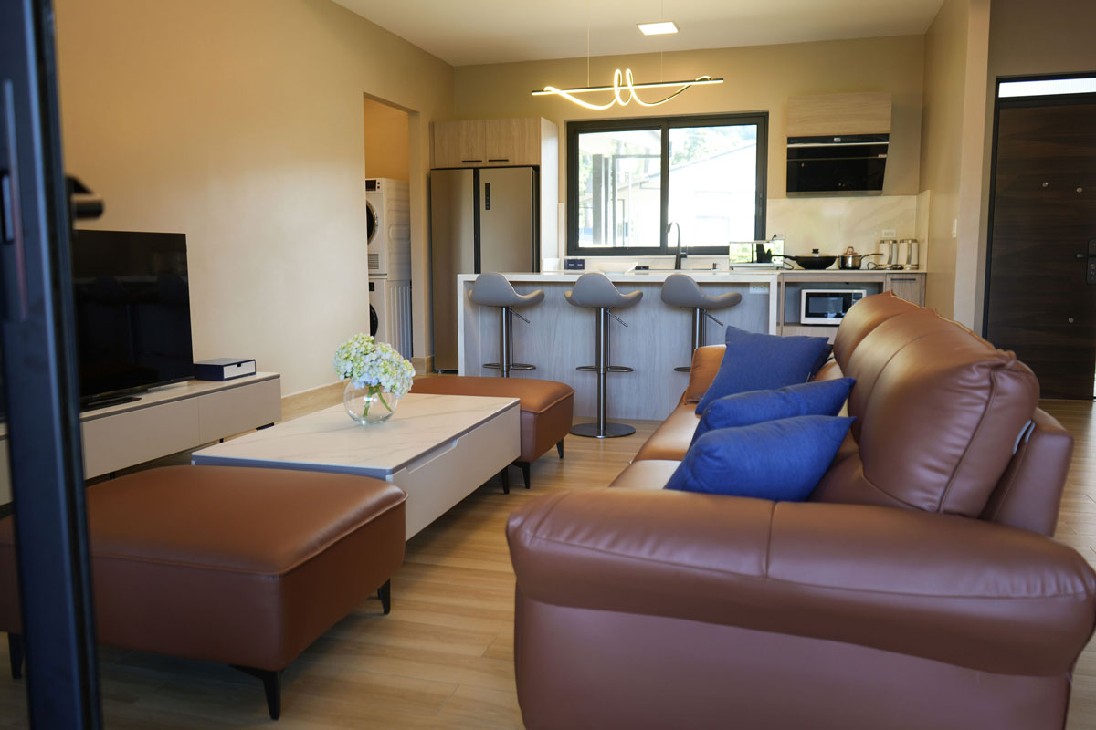 Sala de estar con sofá de cuero marrón, cojines azules, mesa blanca con flores y cocina moderna con barra y taburetes.