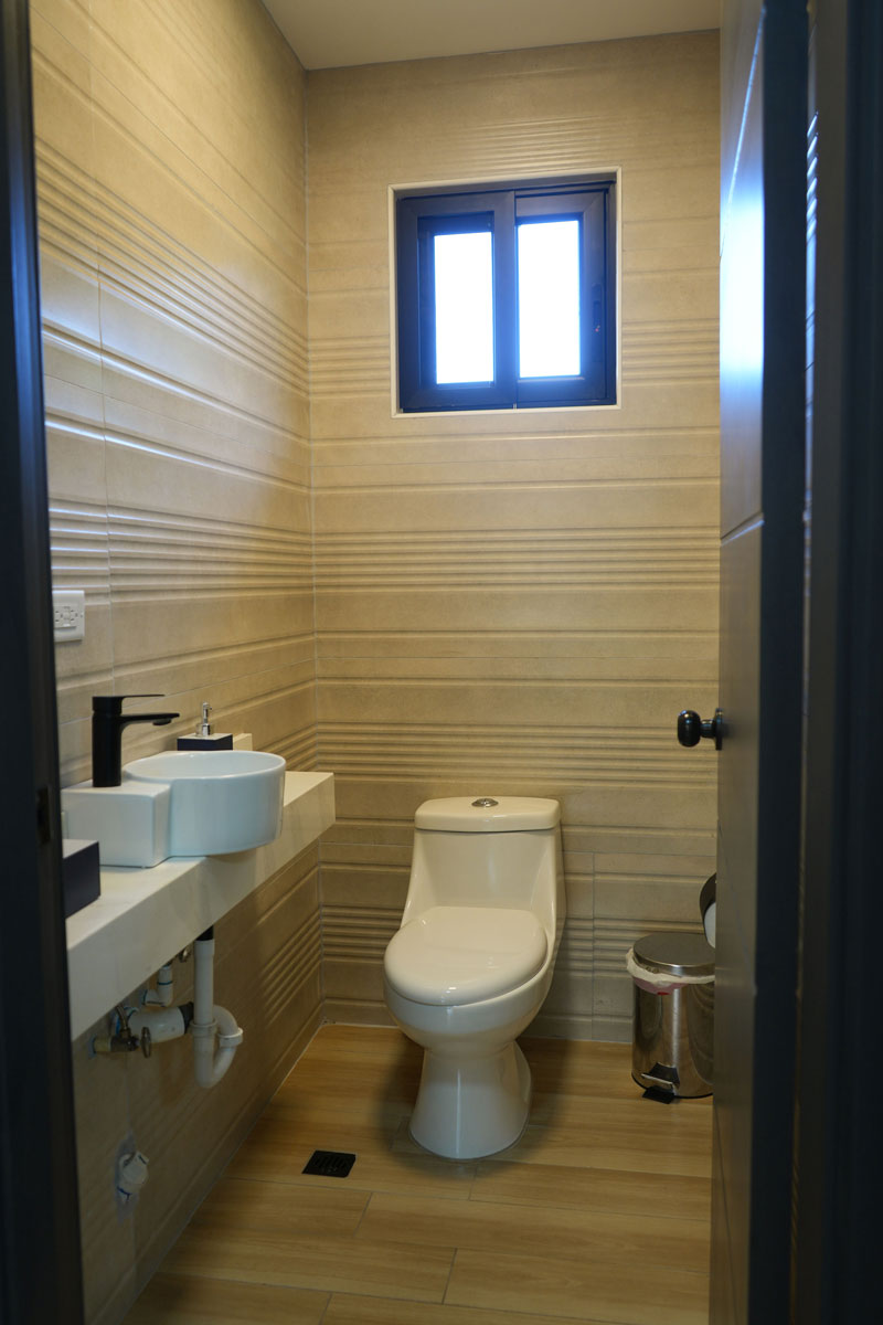 Baño pequeño con inodoro blanco, lavabo con grifo negro, ventana pequeña y bote de basura metálico.