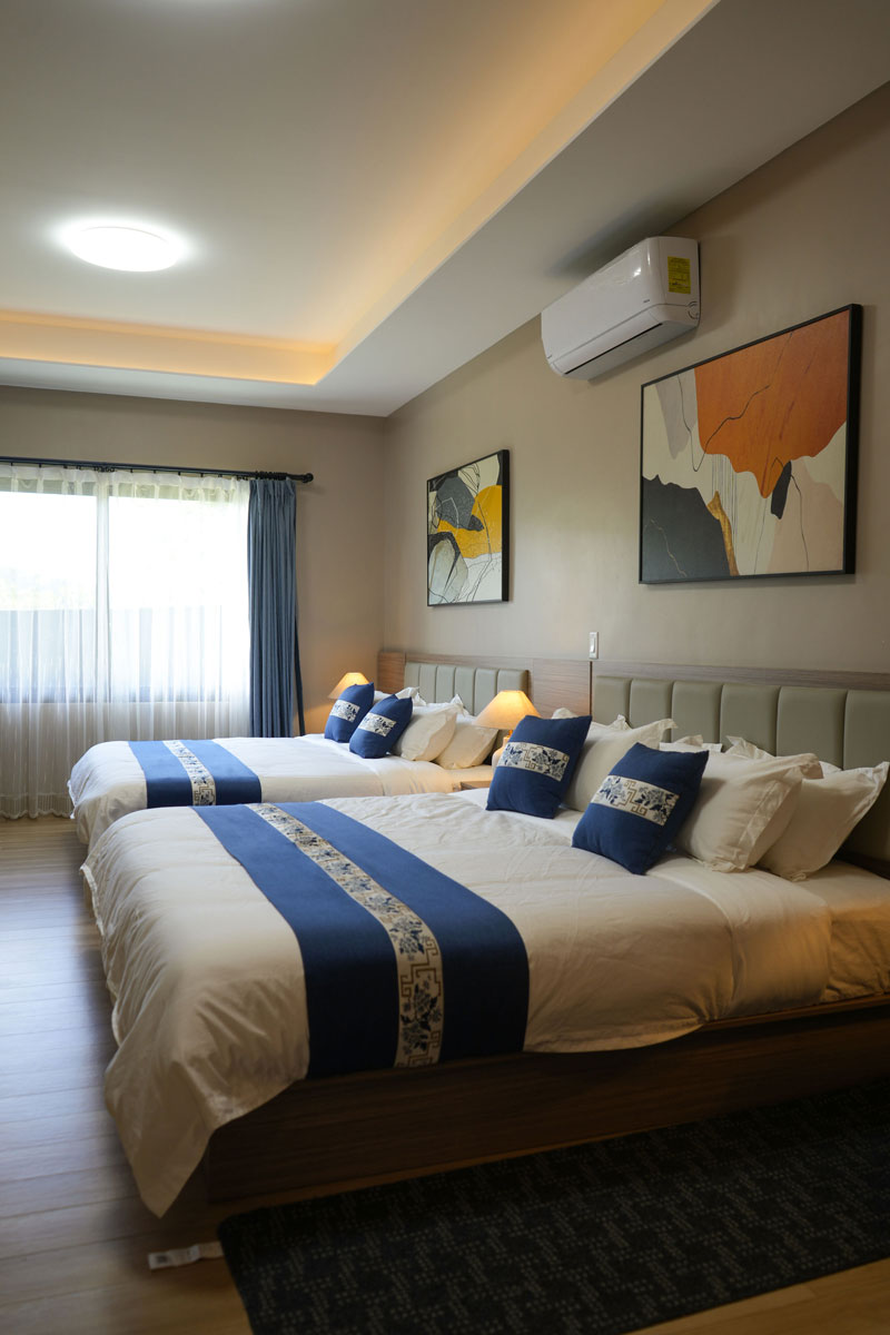 Habitación con dos camas dobles, ropa blanca y decoraciones en azul, ventana con cortinas y dos cuadros abstractos en la pared.