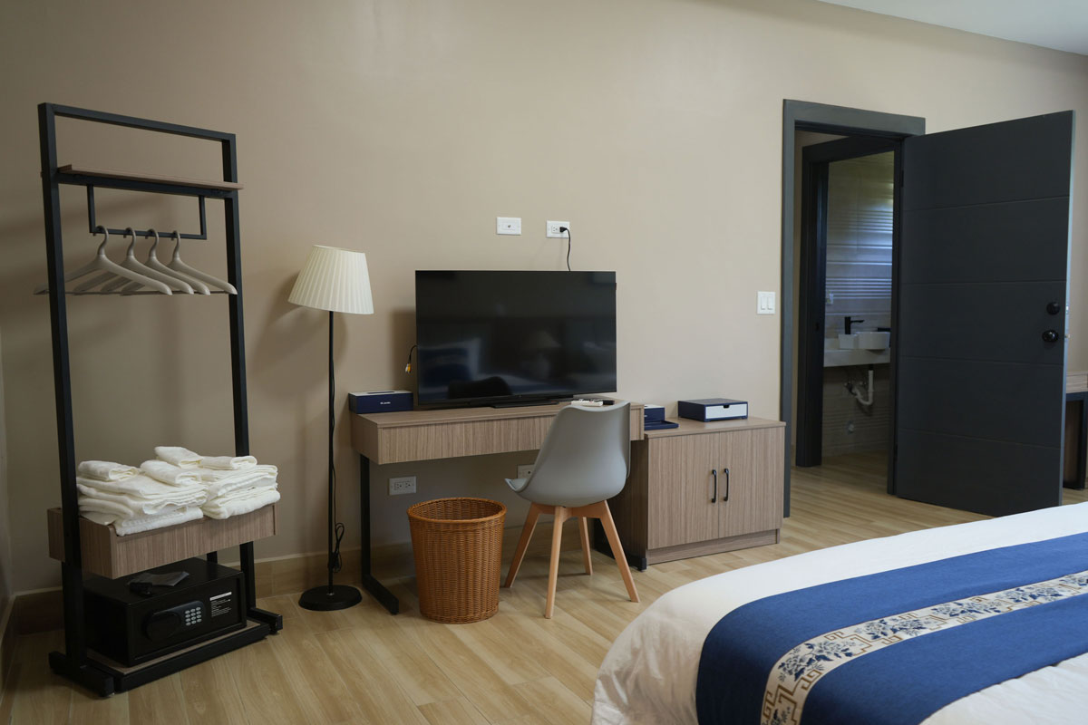 Habitación con cama, televisor sobre escritorio con silla, lámpara de pie, perchero con perchas y toallas, cesta de mimbre y puerta abierta hacia baño.