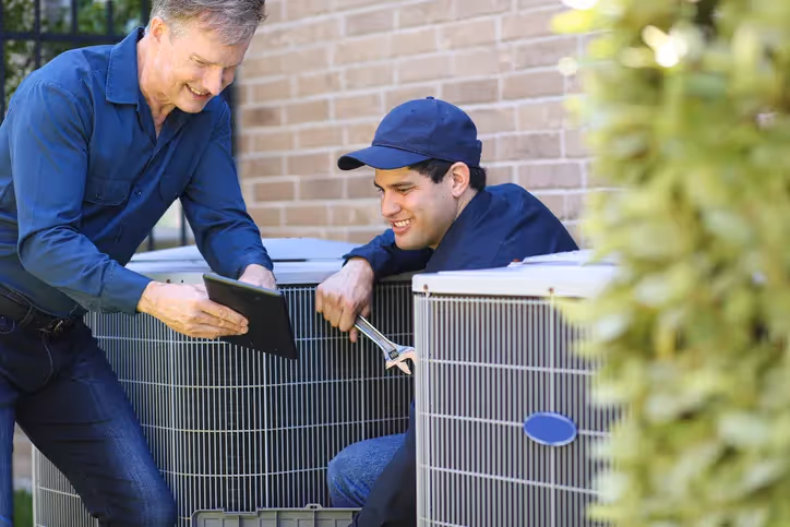 Two HVAC technicians, one with a wrench, review information on a tablet while servicing an outdoor AC unit