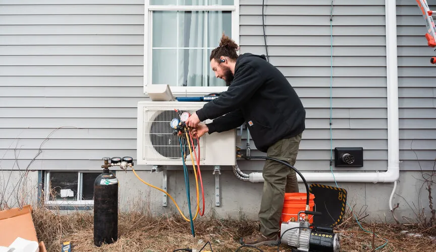 A bearded technician installs an outdoor mini-split heat pump on a gray house, connecting gauges and hoses