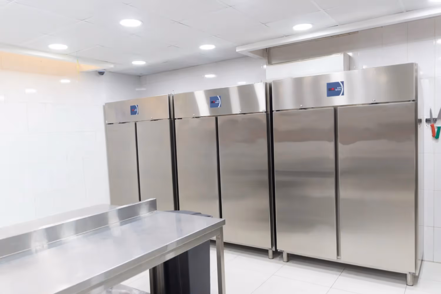 Stainless steel commercial refrigeration units and a prep table in a brightly lit kitchen with white tiled walls.