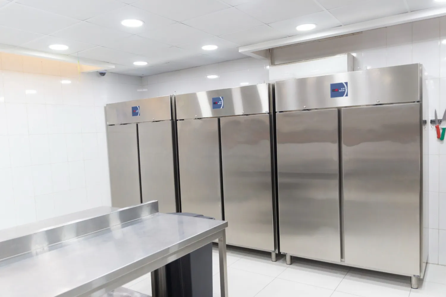 Stainless steel commercial refrigeration units and a prep table in a brightly lit kitchen with white tiled walls.