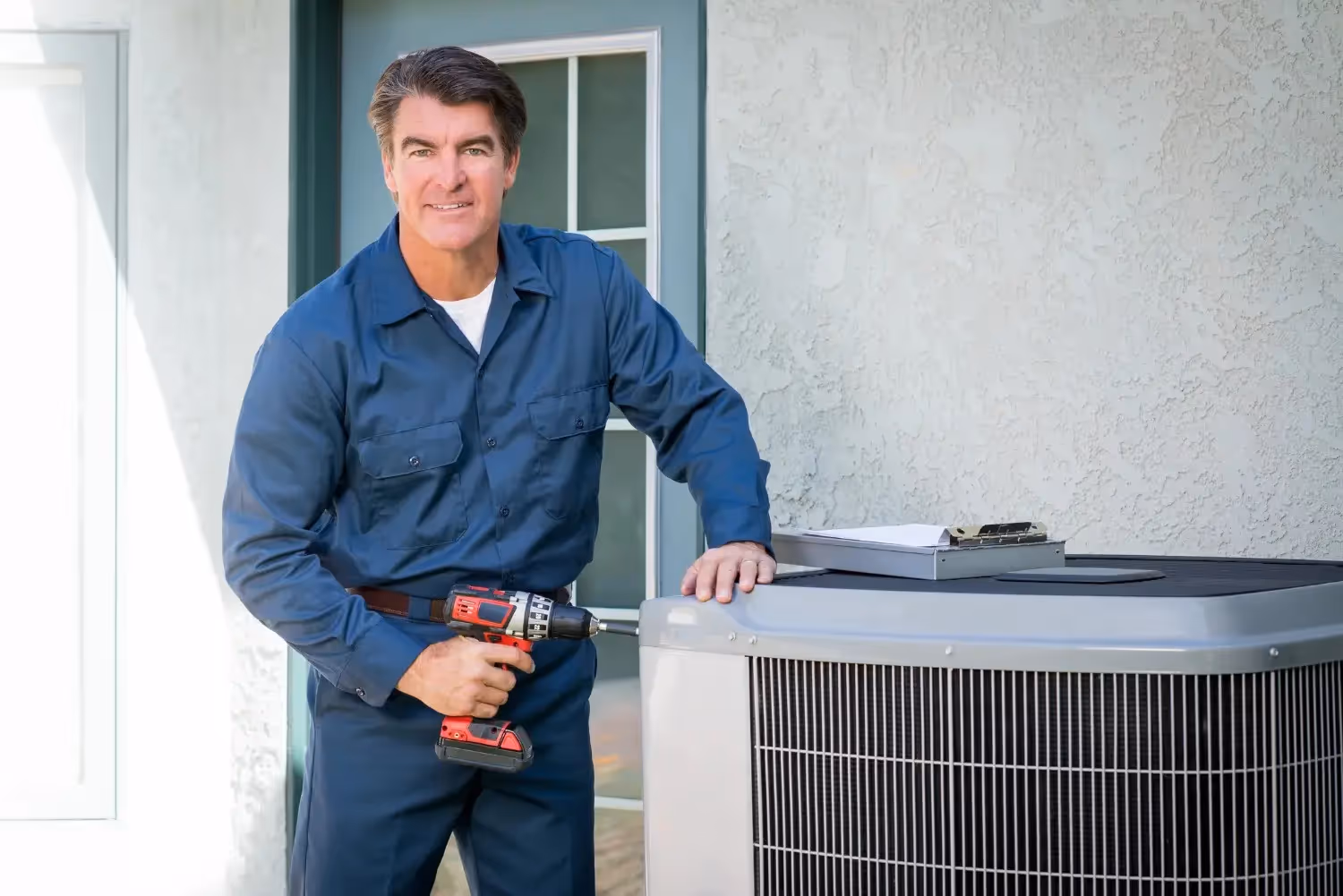 A smiling HVAC technician holding a power drill stands proudly next to a large outdoor air conditioning unit