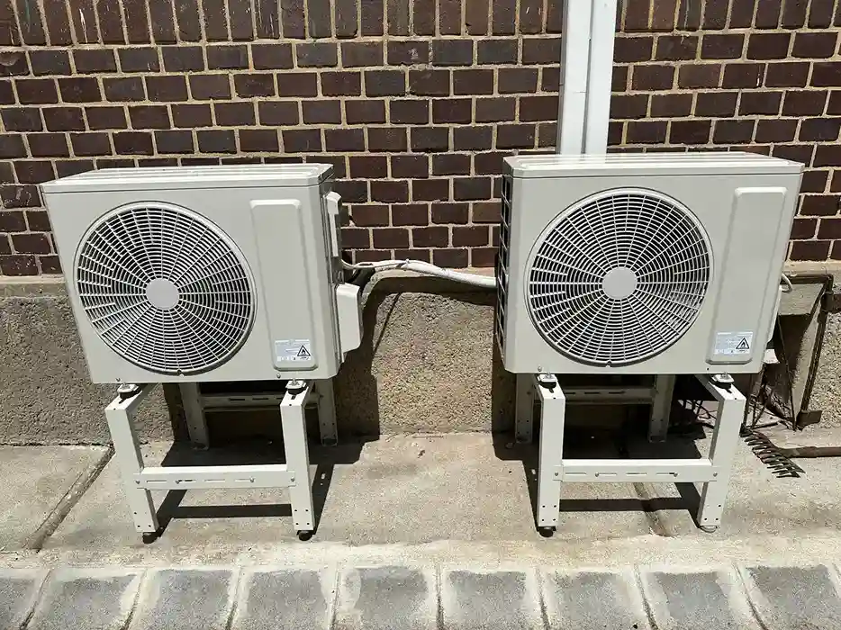 Two white outdoor mini-split air conditioning units on elevated stands against a brick wall.