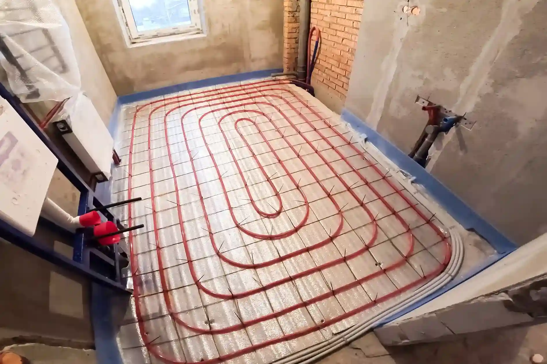 A wide-angle shot of a room under construction, displaying a network of red radiant heating pipes laid on a gray grid on the floor.