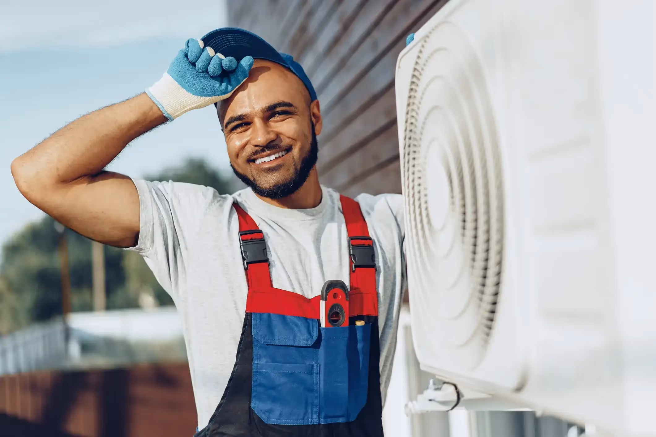 A smiling HVAC technician in overalls and a cap standing next to an outdoor air conditioning unit.