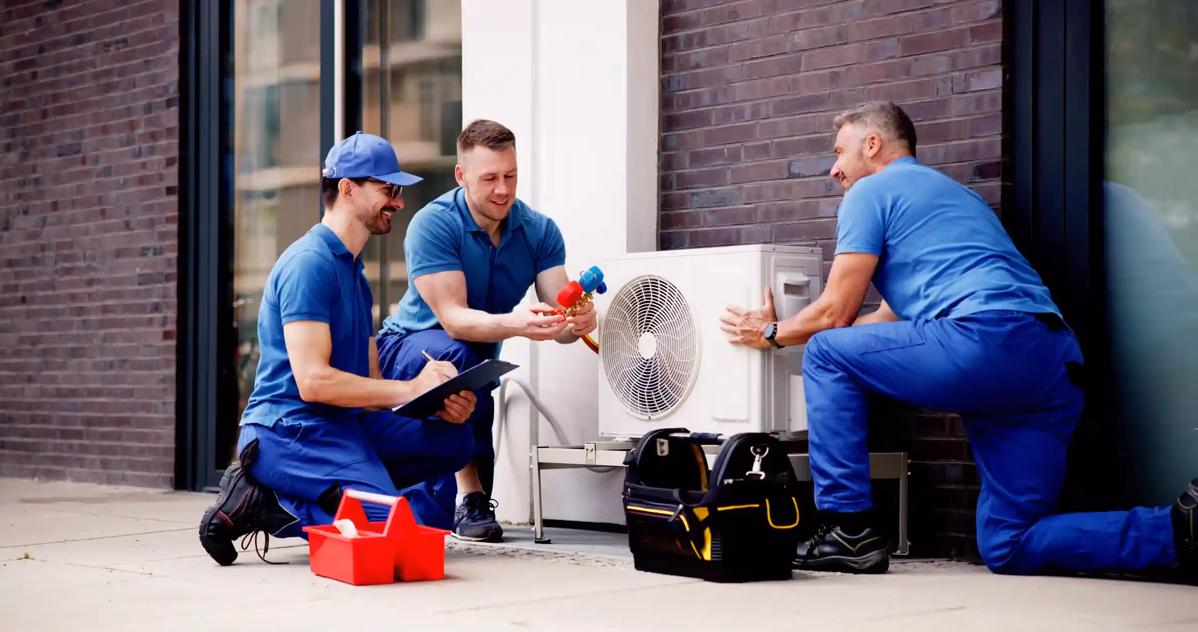 Three male technicians in matching blue shirts and pants are installing an outdoor heat pump unit next to a brick building. One technician is kneeling and holding the unit, another is kneeling and holding manifold gauges, and the third is kneeling while writing on a clipboard. A red toolbox and a black and yellow tool bag are nearby.