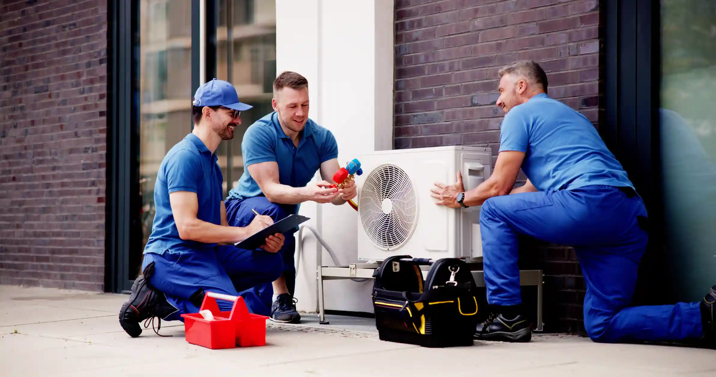 Three male technicians in matching blue shirts and pants are installing an outdoor heat pump unit next to a brick building. One technician is kneeling and holding the unit, another is kneeling and holding manifold gauges, and the third is kneeling while writing on a clipboard. A red toolbox and a black and yellow tool bag are nearby.