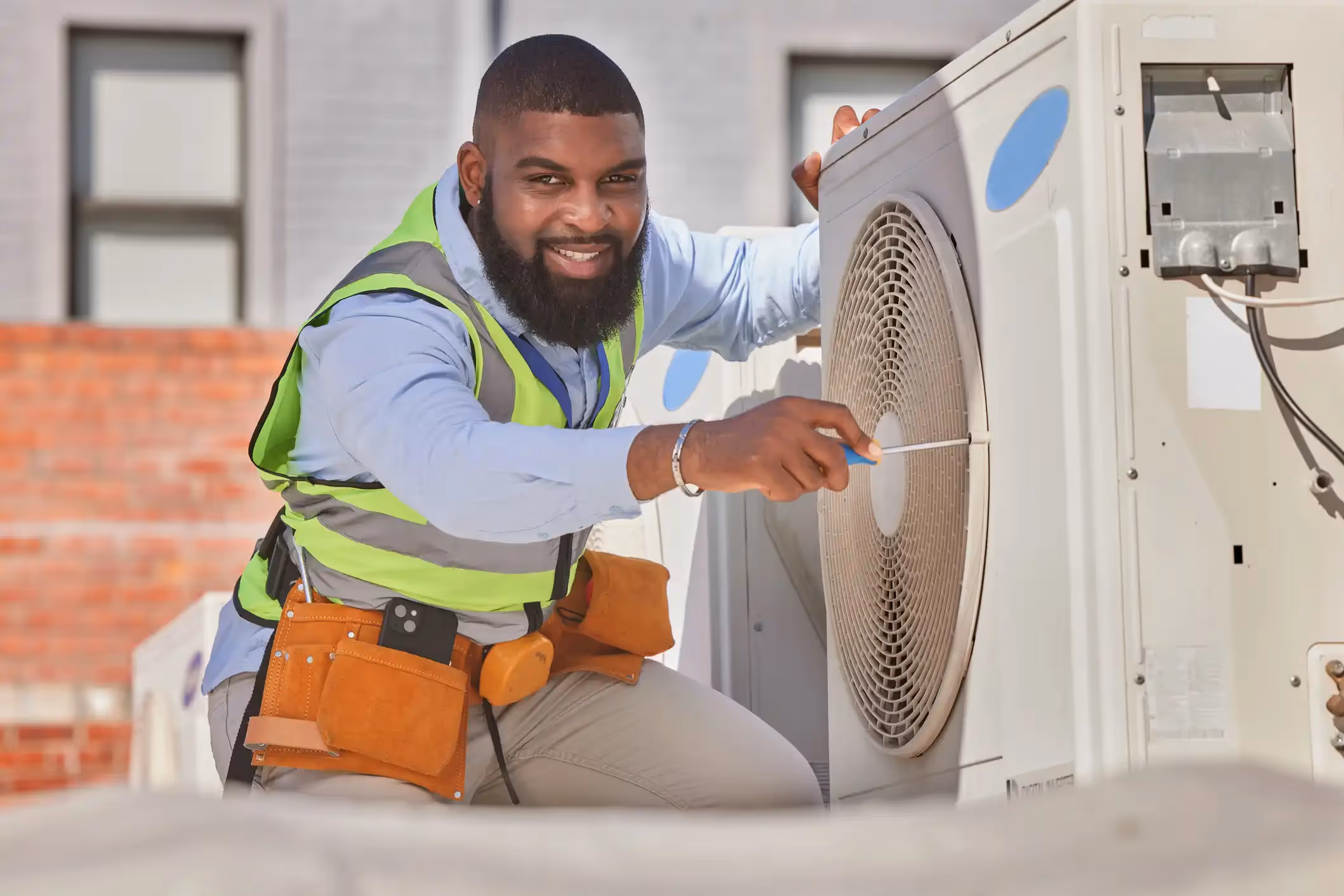 Technician in safety vest adjusts rooftop AC unit fan with screwdriver, tool belt and brick wall visible.