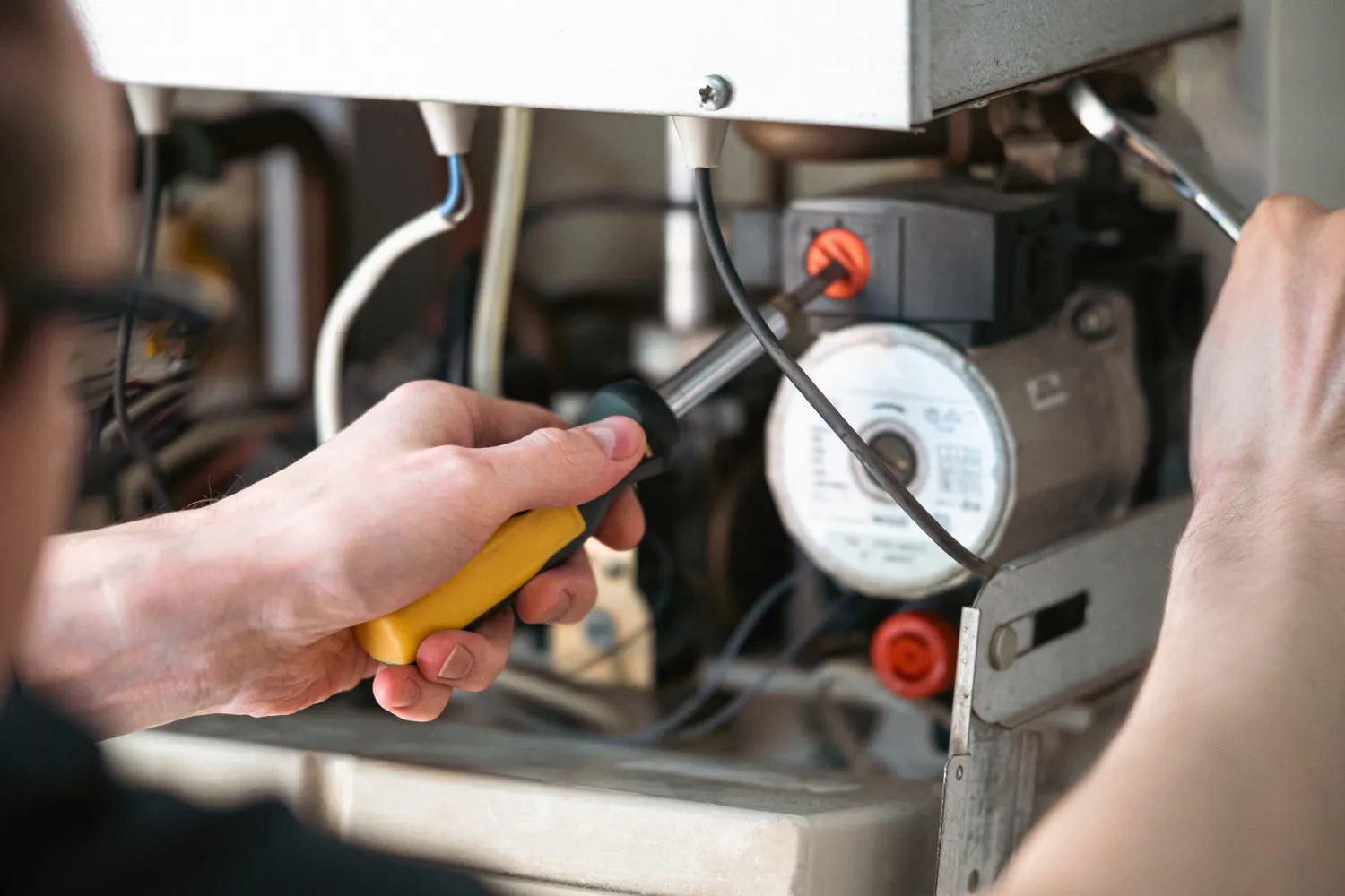 A smiling technician in a dark blue shirt, cap, and tool belt with yellow gloves is using a wrench to work on the top of an outdoor HVAC unit. Another technician, whose back is visible, kneels in the foreground. They are working outside against a brick wall.