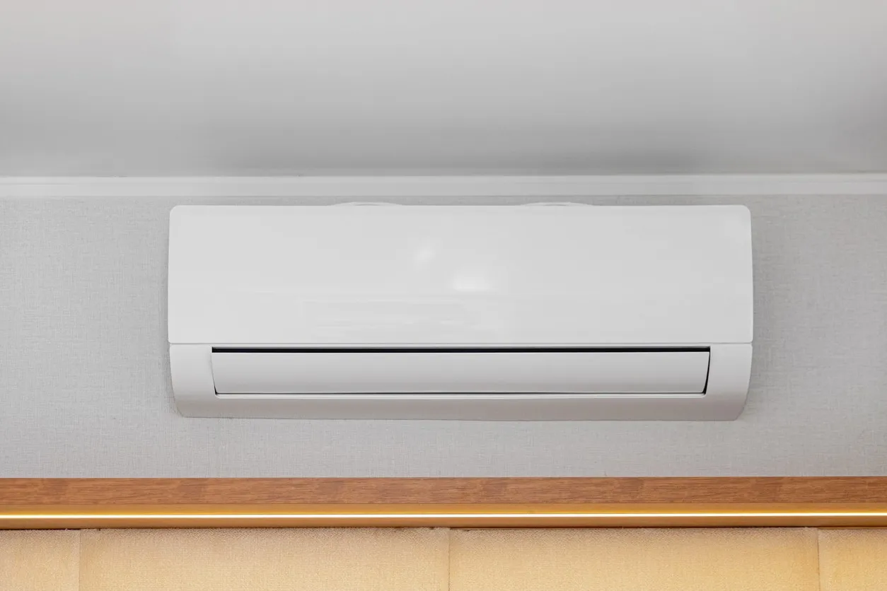 A white ductless mini-split air conditioner indoor unit is mounted on a light gray patterned wall. The unit is closed and appears to be functioning, with the air vent visible at the bottom. It is installed high up, near a white ceiling, above a wooden trim and a light-colored, upholstered headboard or panel.