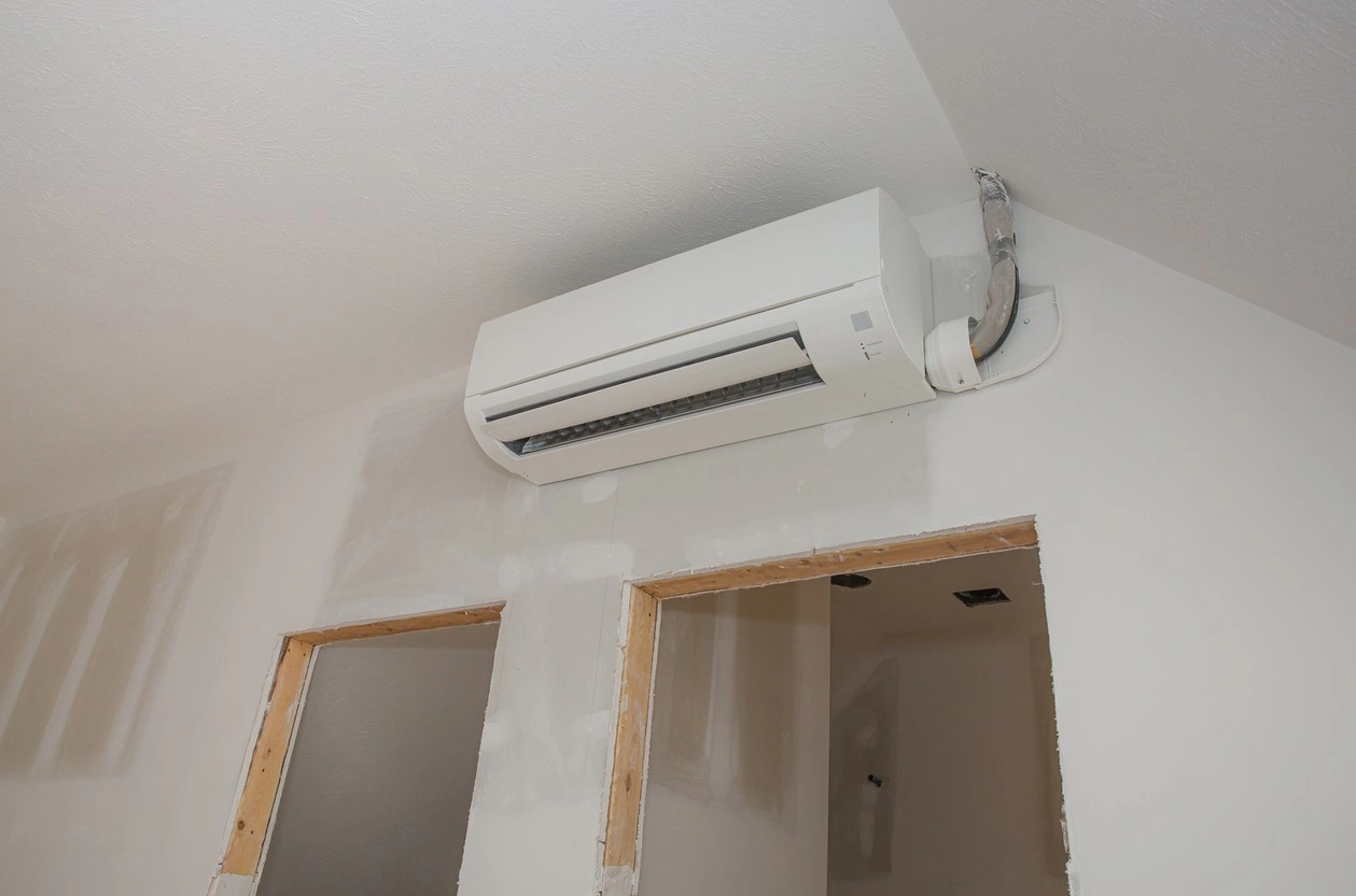 A white ductless mini-split air conditioner indoor unit is mounted on a pale wall with a vaulted ceiling, likely in a room under construction or renovation, as the walls are rough and unpainted. The insulated refrigerant lines and drainage pipe exit the unit's right side and curve upwards toward the ceiling. There are two rough door openings beneath the unit.