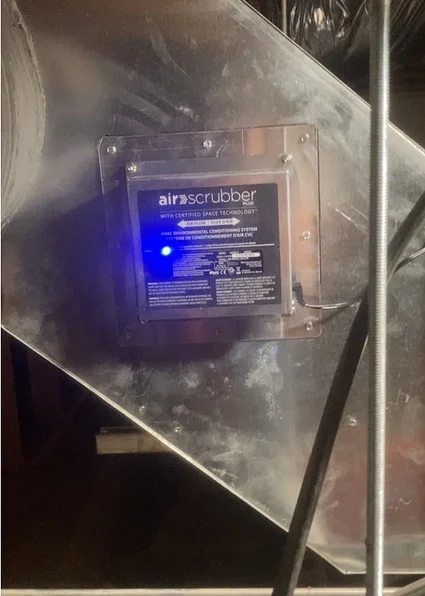 Air scrubber device installed in ductwork, featuring a blue indicator light, highlighting air purification technology for improved indoor air quality in homes.