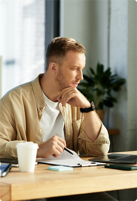 Man in beige shirt studying notes while sitting at a desk with a laptop and coffee cup.