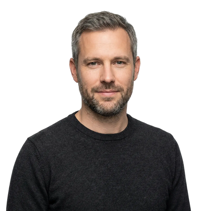 Portrait of a smiling middle-aged man with short gray hair and beard wearing a dark crewneck sweater against a transparent background.