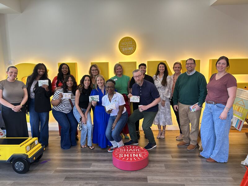 A group of people are gathered around tables, participating in Mind Above Matter's 2025 St. Patty's Day Affirmation Box Workshop, co-sponsored by The Maples and Meridell Achievement Center, in Grapevine, TX. The participants are engaged in creating affirmation boxes, with various materials and supplies visible on the tables. Banners in the background read "HOUSE," "SHINE," and "SHARE YOUR SHINE."