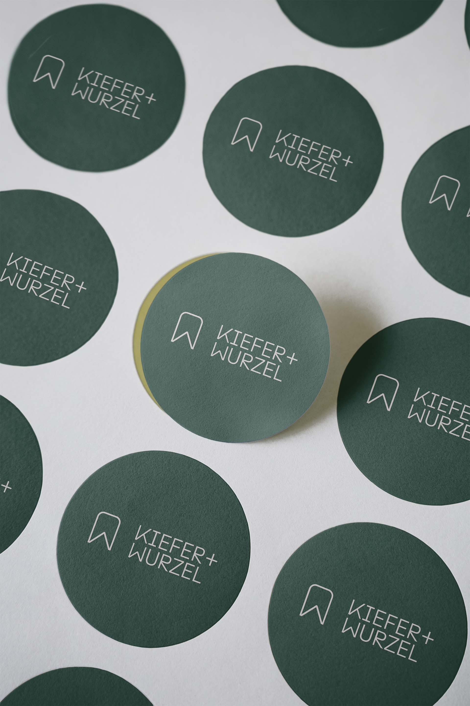 Mehrere grüne runde Aufkleber mit weißem Schriftzug KIEFER+WURZEL und einem Lesezeichen-Symbol auf weißem Hintergrund.