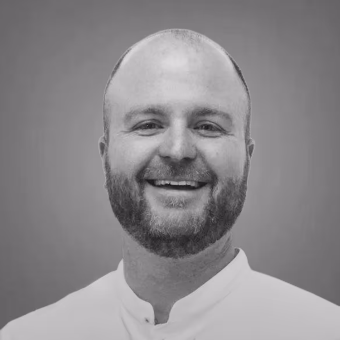 Smiling bald man with a beard wearing a white collared shirt against a plain background.