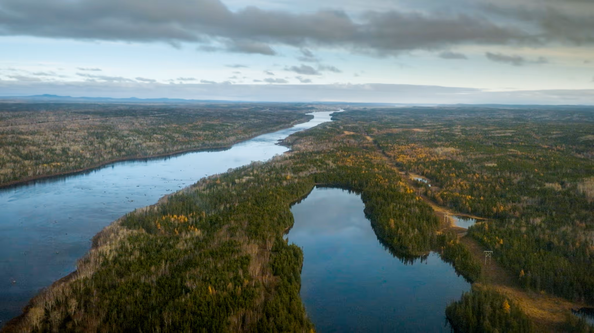 Aerial view of a winding river flowing through a dense forest with some areas of autumn foliage under a cloudy sky.