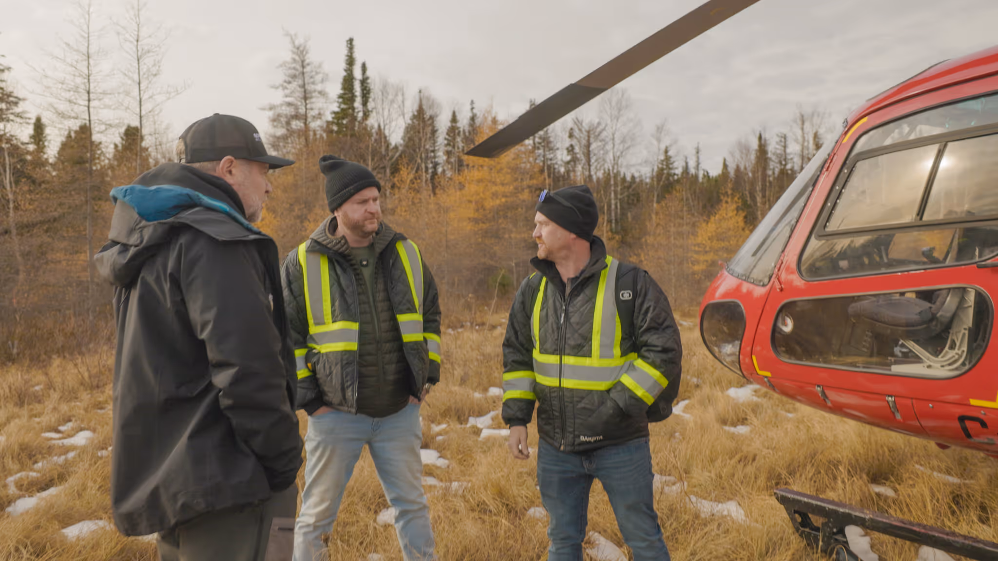 Three men wearing jackets and reflective vests standing near a red helicopter on a grassy field with patches of snow and autumn trees in the background.