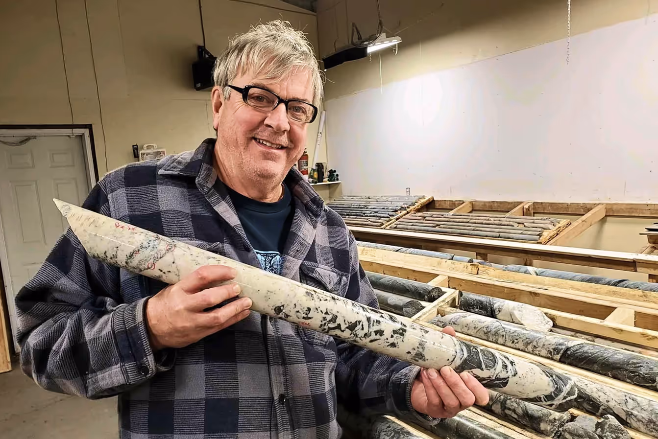 Smiling man in glasses and plaid shirt holding a large cylindrical rock core sample inside a lab with more core samples on wooden racks.