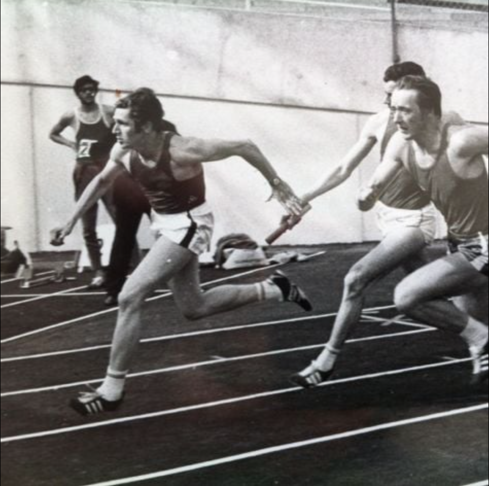 Course de relais en salle avec passage de témoin entre deux athlètes sur une piste marquée.