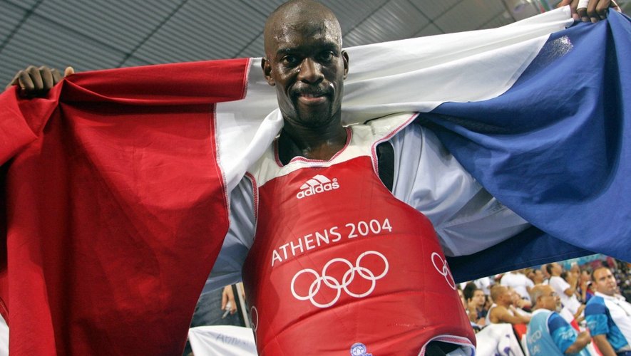 Athlète tenant un drapeau français, portant une tenue rouge avec le logo et l'inscription Athens 2004 des Jeux Olympiques.