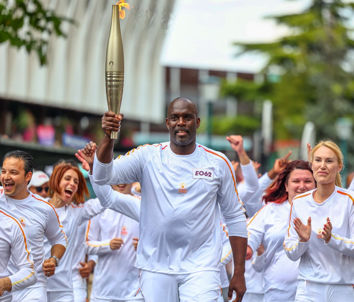 Un homme tenant une torche olympique en flammes, entouré de personnes en tenues blanches célébrant dans une ambiance joyeuse en extérieur.