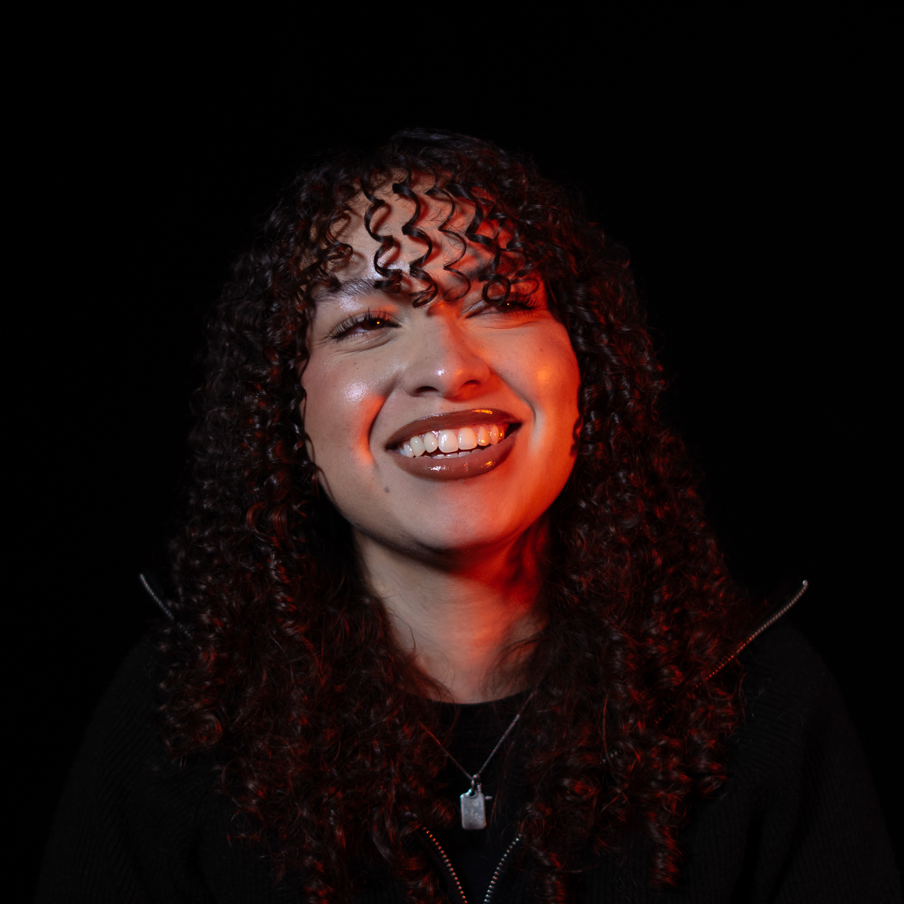 Jeune femme souriante aux cheveux bouclés portant un pendentif rectangulaire sur fond noir.