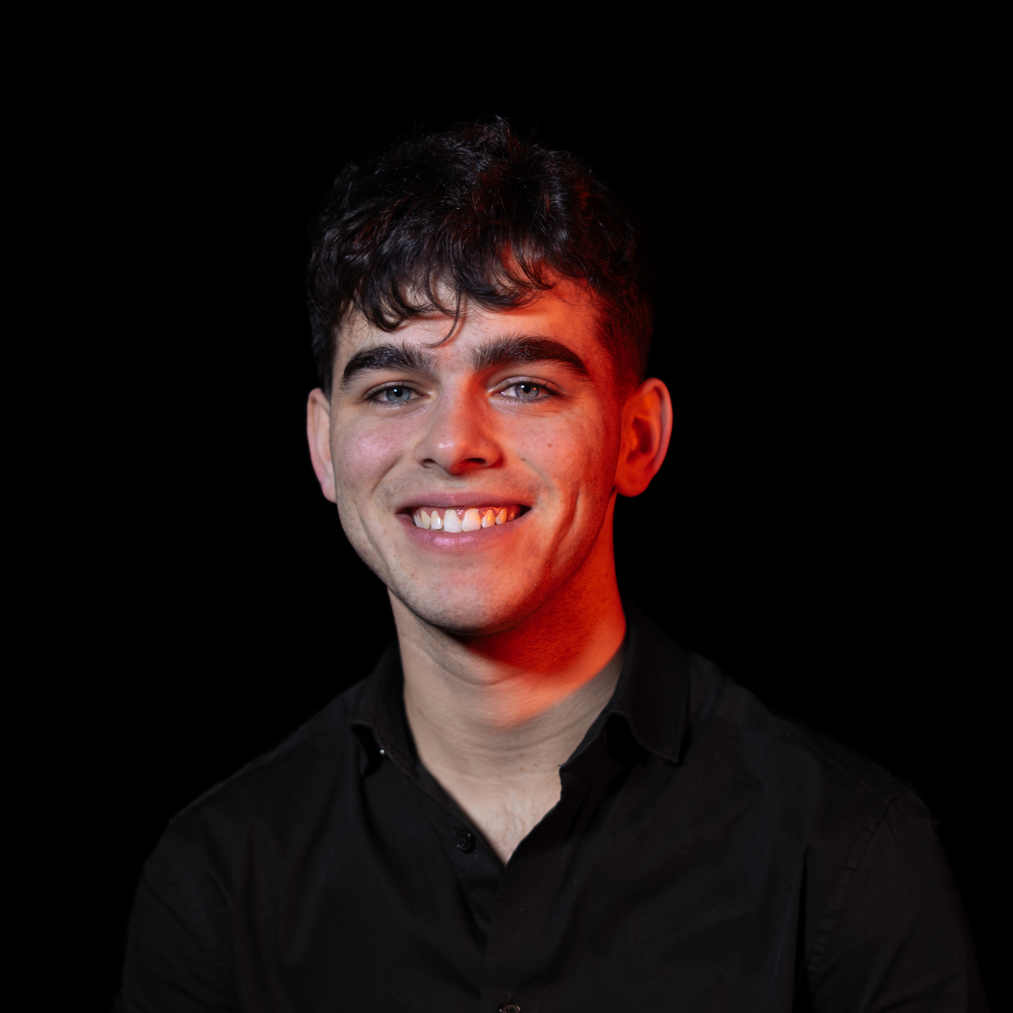 Jeune homme souriant avec cheveux bouclés courts et chemise noire sur fond noir, éclairé par une lumière rouge latérale.