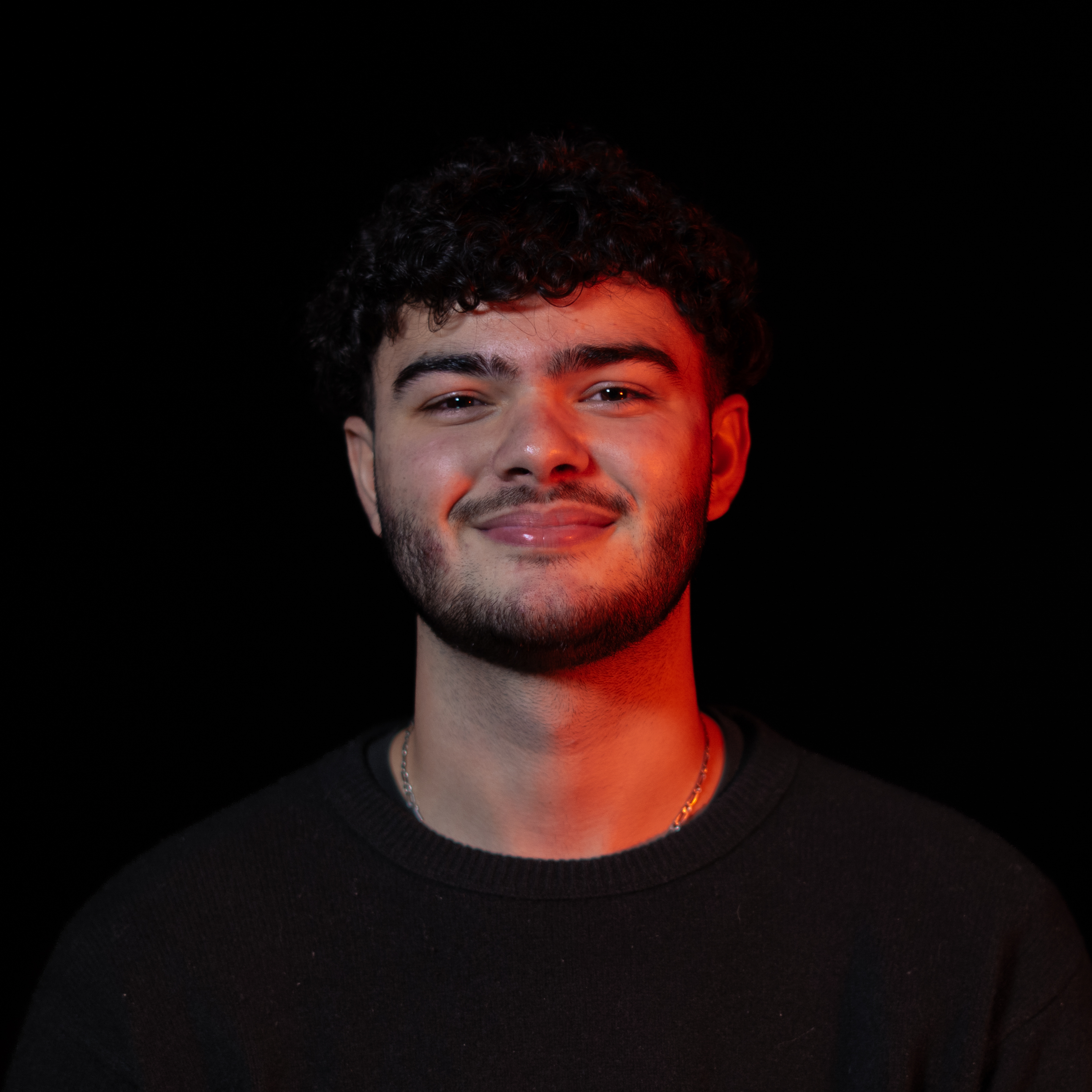 Portrait d'un jeune homme souriant avec des cheveux bouclés et une barbe, éclairé d'une lumière rouge sur fond noir.