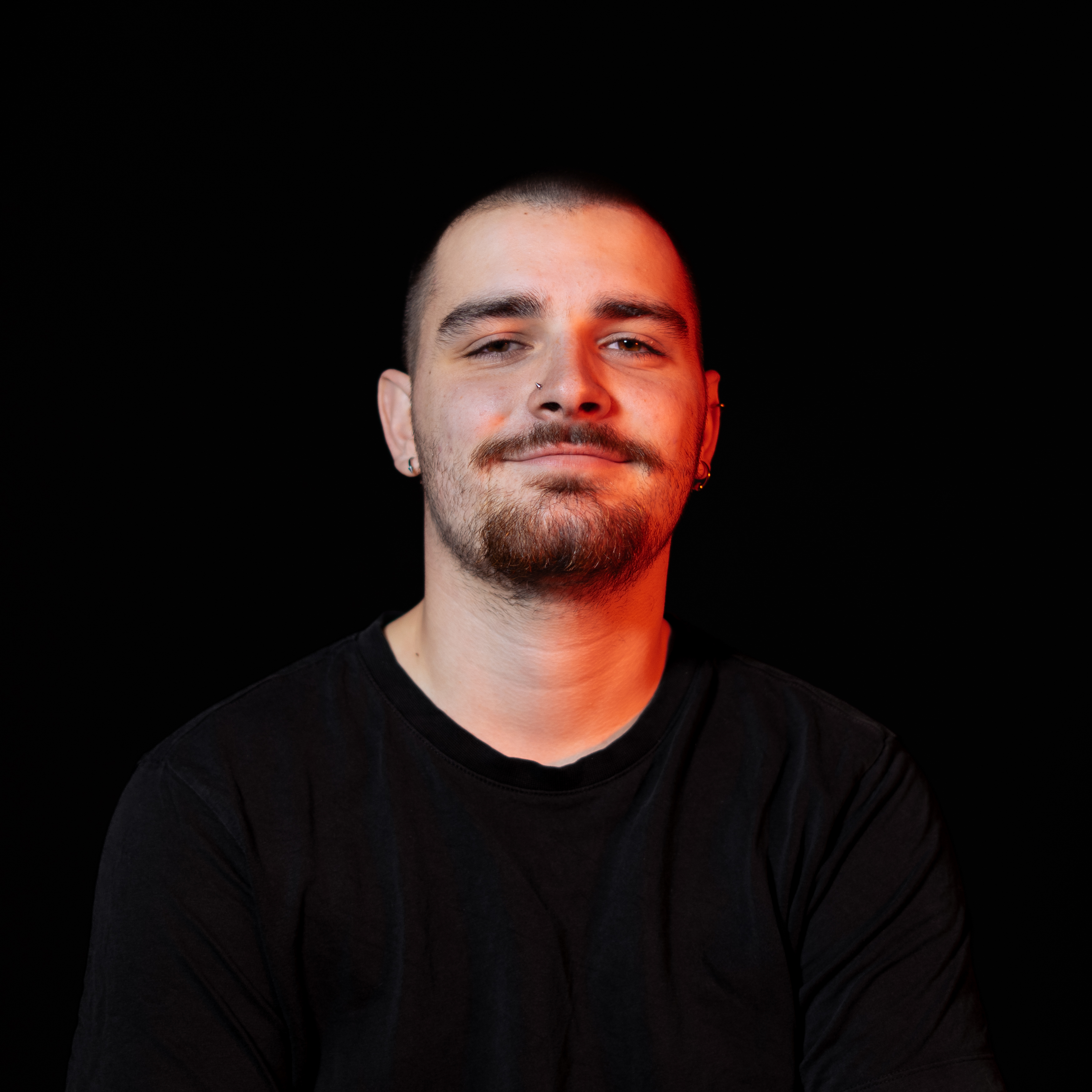 Homme souriant avec barbe et piercings, portant un t-shirt noir sur fond noir.