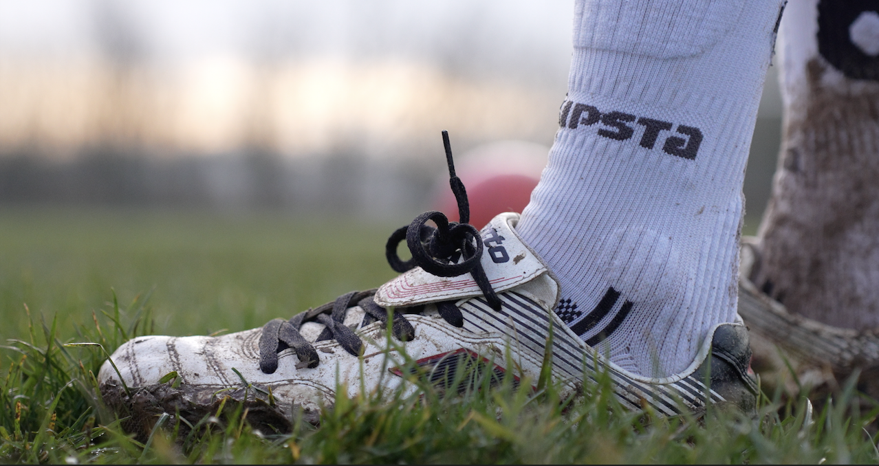 Chaussure de football blanc avec lacets noirs et chaussettes blanches sur de l'herbe avec de la boue.