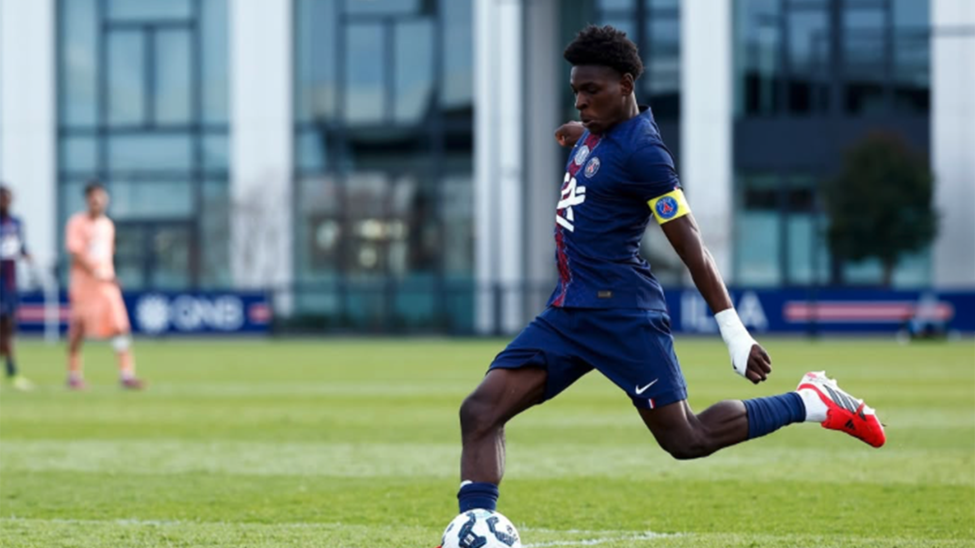 Joueur de football de l'équipe PSG en train de tirer un ballon sur un terrain avec un bâtiment moderne en arrière-plan.