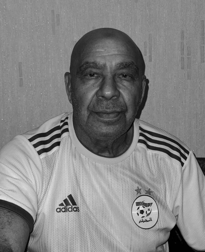 Homme âgé chauve portant un maillot de football Adidas de l'équipe algérienne, regardant vers l'avant.