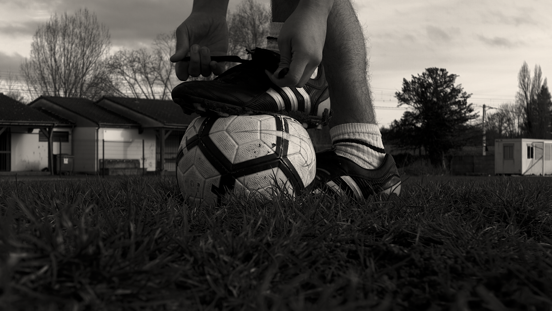 Joueur de football attachant ses chaussures avec un pied posé sur un ballon sur un terrain herbeux.