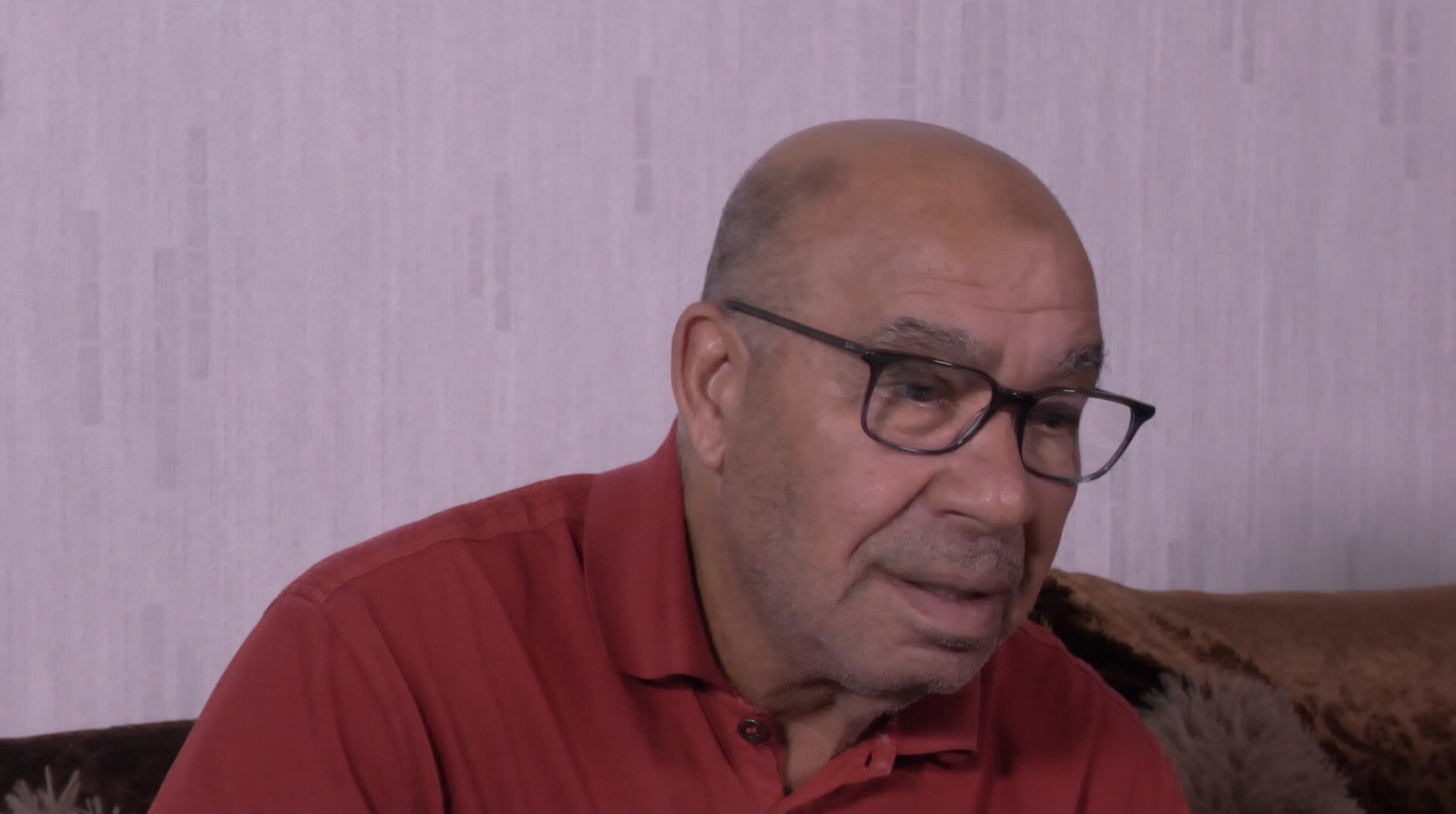 Homme âgé portant des lunettes et un polo rouge, assis devant un mur clair.