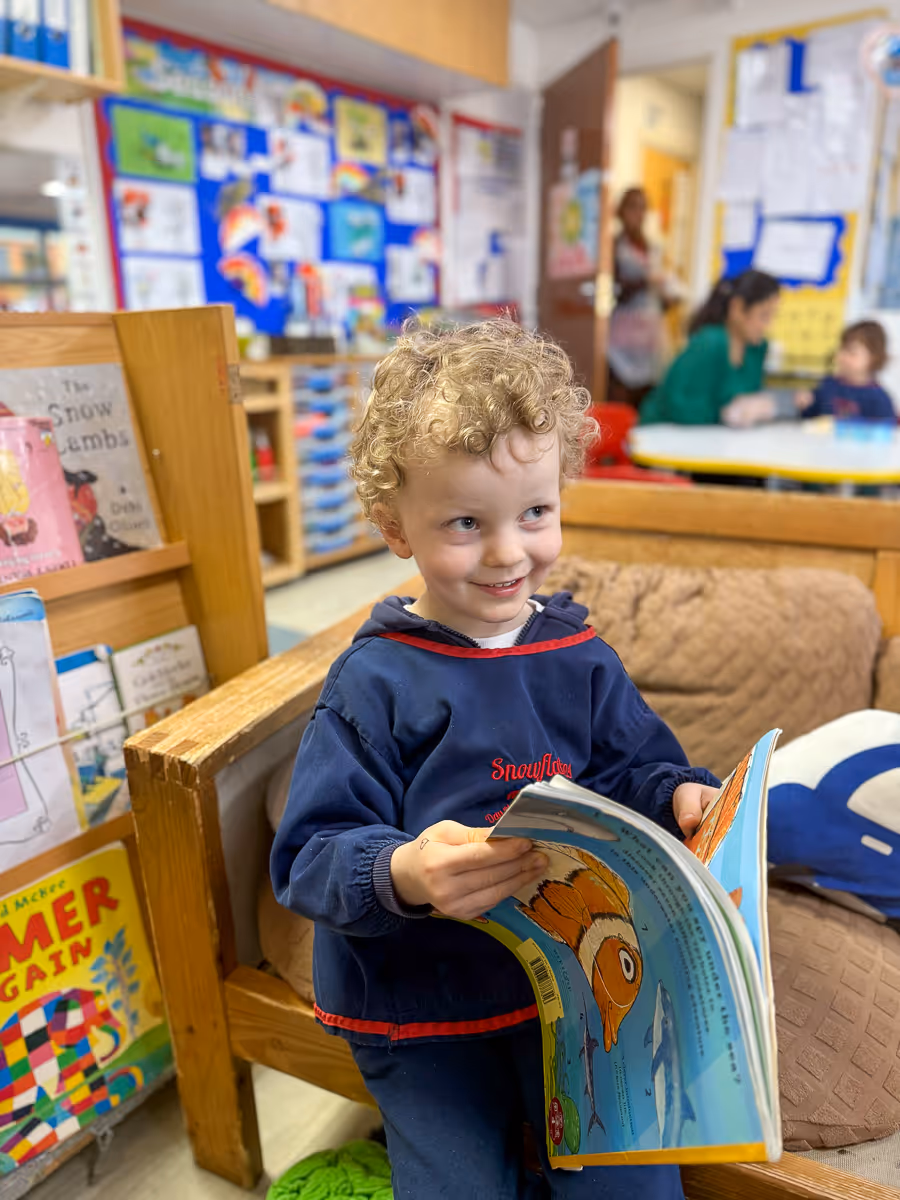 Smiling young boy with curly blonde hair holding and looking at an open colorful fish-themed book in a classroom reading area.