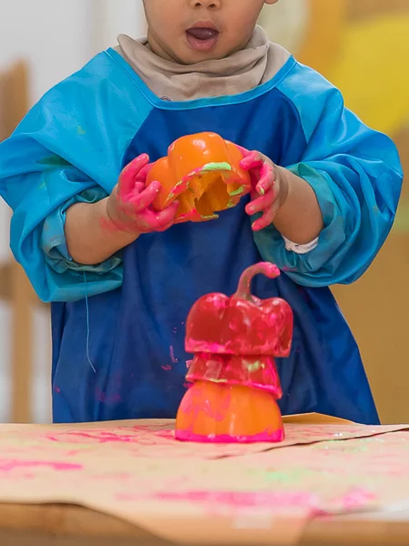 Child wearing a blue smock playing with painted orange bell peppers, hands covered in pink paint.